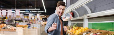 Cheerful man pointing with fingers near fruits in supermarket, banner