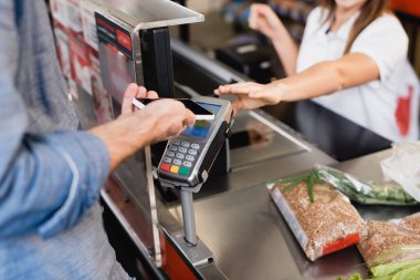 Markette arka plandaki bulanık kasiyer ve bakkaliye yakınlarında akıllı telefonla ödeme yapan bir adamın görüntüsü. 