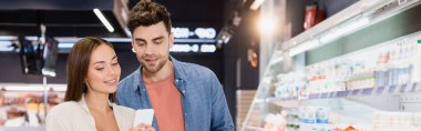 Young couple using smartphone in supermarket, banner 
