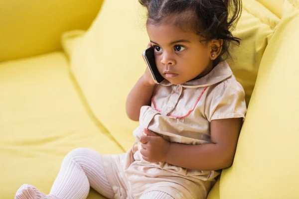 幼いアフリカ系アメリカ人の女の子がソファに座っている間にスマートフォンで話しています — ストック写真