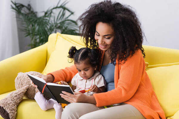 smiling african american mother sitting with toddler daughter and looking at notebook in living room