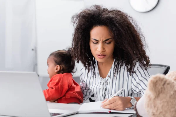 stressed african american mother holding in arms crying toddler daughter while working from home