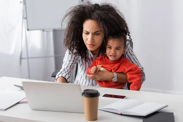 stressed african american mother holding in arms crying toddler kid while working from home