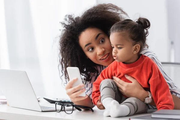 アフリカ系アメリカ人の母親が机の上で娘にスマートフォンを見せ — ストック写真