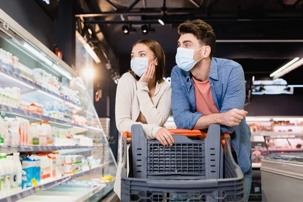 Casal Máscaras Médicas Olhando Para Mantimentos Perto Carrinho Compras Supermercado — Fotografia de Stock