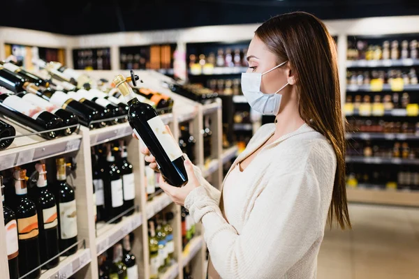 Kvinna Medicinsk Mask Tittar Vin Flaska Nära Hyllor Suddig Bakgrund — Stockfoto