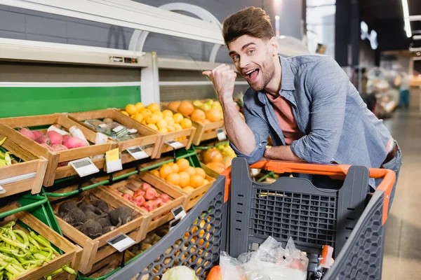 Fröhlicher Mann Zeigt Auf Lebensmittel Auf Theke Neben Einkaufswagen Supermarkt — Stockfoto