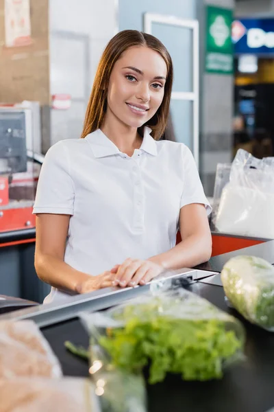 Smiling cashier looking at camera near food on supermarket checkout on blurred foreground