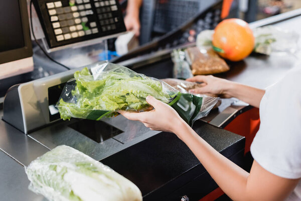Cropped view of young cashier holding fresh lettuce on supermarket checkout