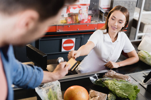 Smiling cashier taking credit card from customer rear groceries on checkout in supermarket 