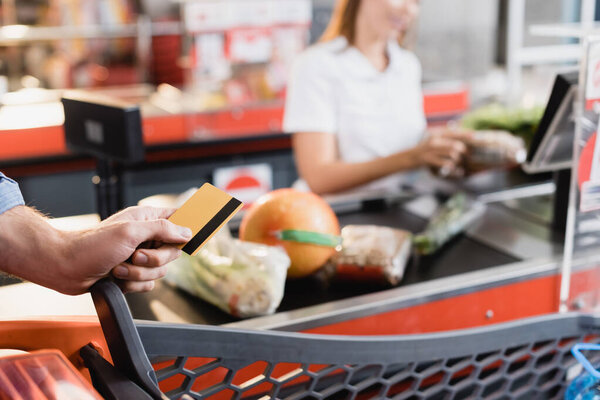 Cropped view of man holding credit card near shopping cart and supermarket checkout on blurred background 
