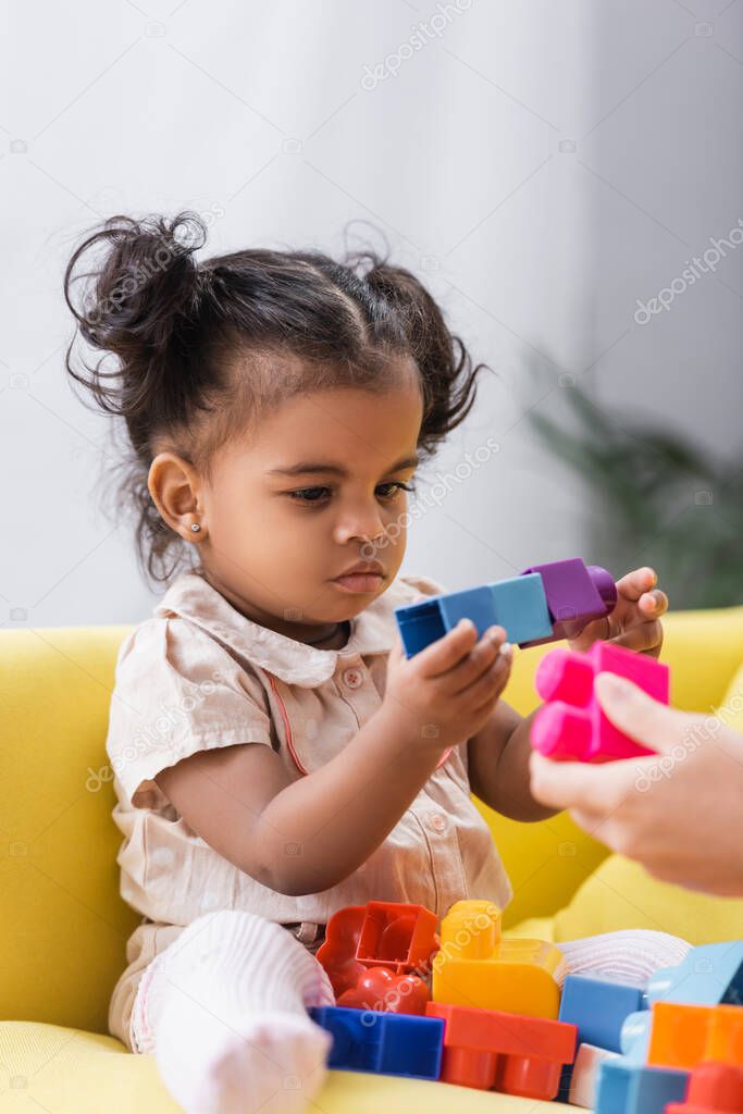 african american toddler girl sitting on couch and playing building blocks with mother on blurred foreground