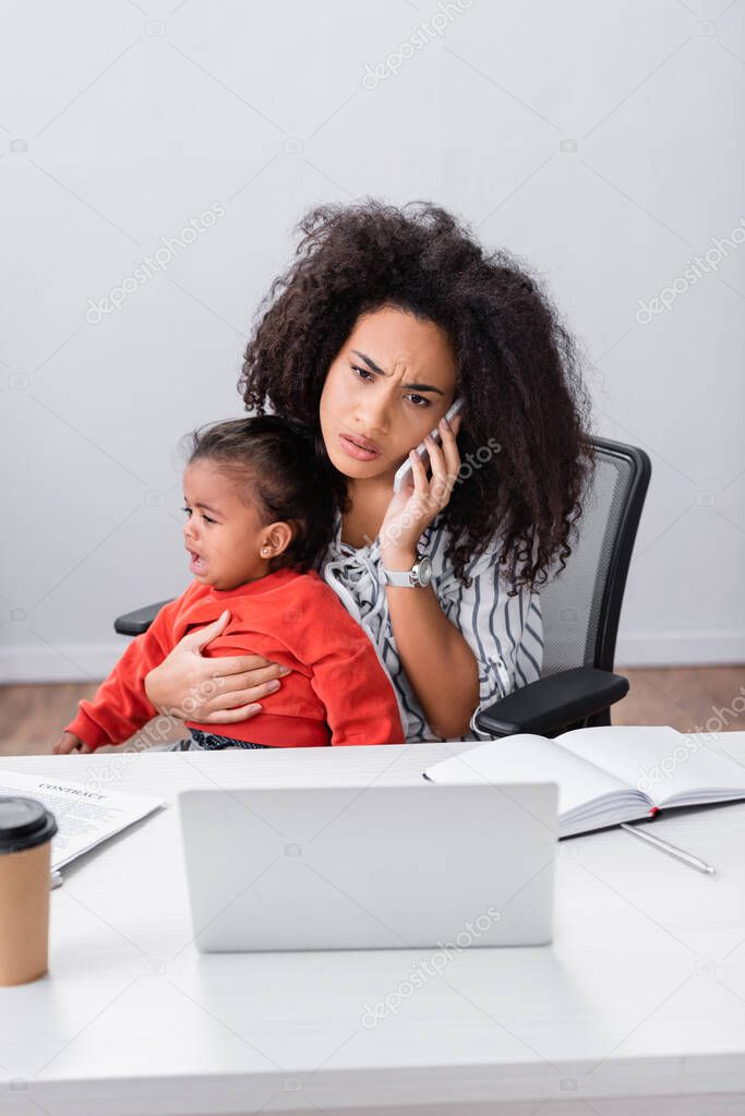 stressed african american freelancer holding in arms crying toddler daughter while talking on smartphone 