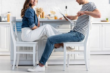 Sandalyeye oturmuş neşeli bir kadının yanında gazete okuyan bir adam. 