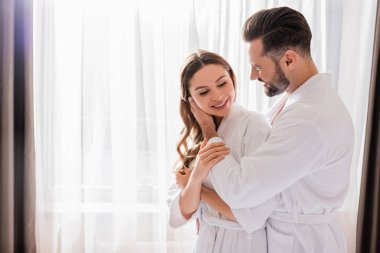 Bornozlu bir adam otel odasının penceresinin yanında gülümseyen kız arkadaşının yüzüne dokunuyor. 