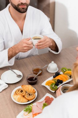 Cropped view of man in bathrobe holding cup of coffee near tasty breakfast on table and girlfriend on blurred foreground in hotel  clipart