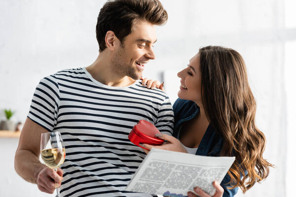 man holding glass of wine and newspaper and looking at cheerful girlfriend with heart-shaped gift box