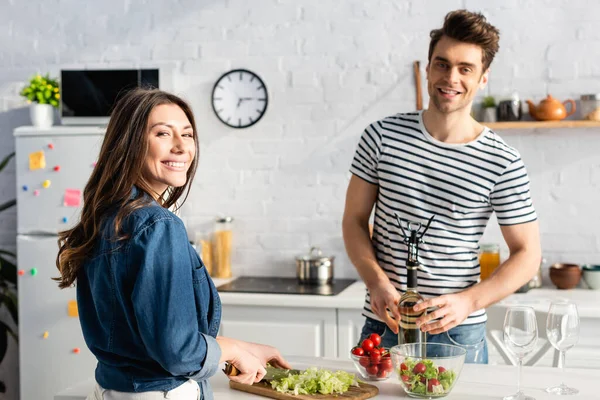 Cheerful Woman Looking Camera While Cutting Lettuce Boyfriend Opening Bottle — Stock Photo, Image