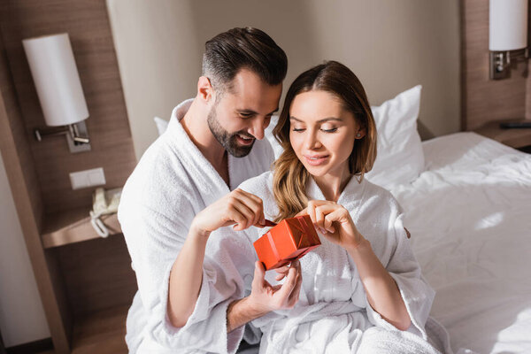 Smiling couple in bathrobes holding present on hotel bed 