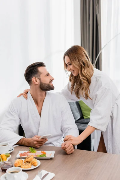 在酒店里 一个拿着智能手机笑着看着穿着浴衣的女朋友的男人 早餐时喝着咖啡 前景暗淡 — 图库照片