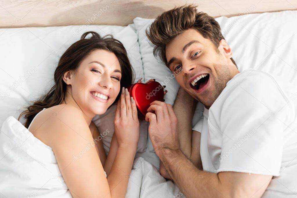 top view of happy couple lying in bed near red heart-shaped box on valentines day