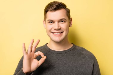 Young man showing okay gesture and looking at camera on yellow background clipart