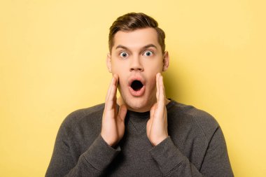 Amazed man with hands near mouth looking at camera on yellow background clipart