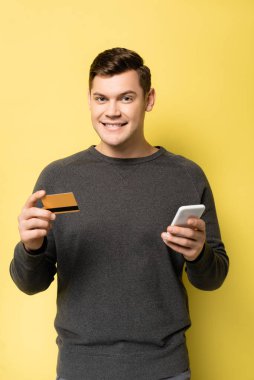 Sarı arka plandaki kameraya bakarken elinde kredi kartı ve akıllı telefon tutan gülümseyen adam.