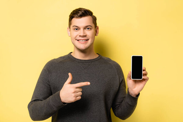 Smiling man pointing with finger at smartphone with blank screen on yellow background