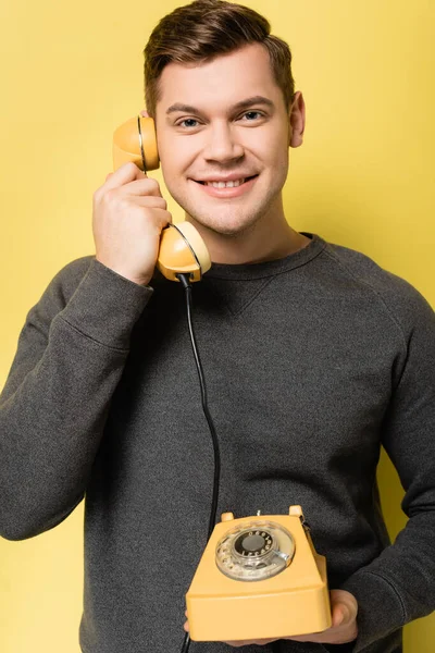 微笑的男人一边看着相机 一边用黄色背景的老式电话交谈 — 图库照片