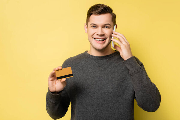 Smiling man talking on cellphone and holding credit card on yellow background