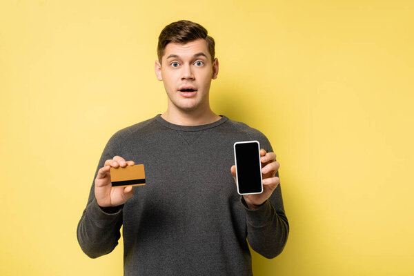 Amazed man holding credit card and smartphone on yellow background