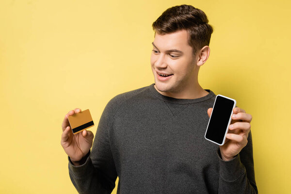 Cheerful man looking at credit card while holding cellphone on yellow background