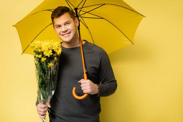 带着花和伞微笑的男人 看着黄色背景的相机 — 图库照片