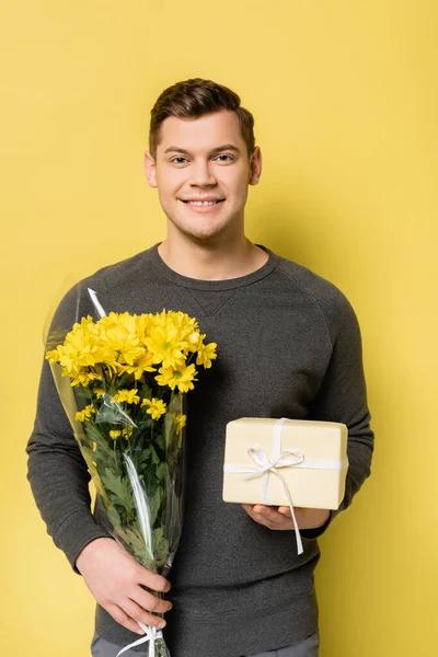 Молодий Чоловік Посміхається Камеру Тримаючи Подарункову Коробку Букет Жовтому Тлі — стокове фото