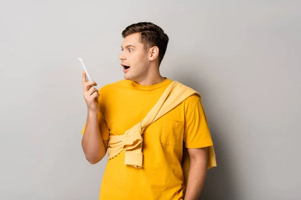 Opgewonden Man Geel Shirt Kijkend Naar Mobiele Telefoon Grijze Achtergrond — Stockfoto