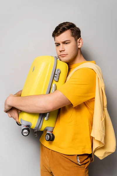 不满的男人抱着灰色背景的黄色行李箱 — 图库照片