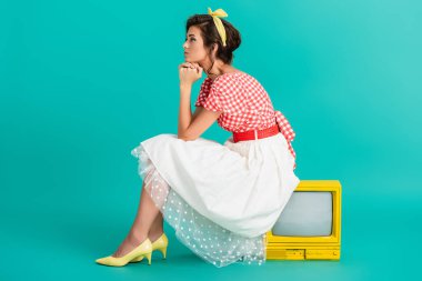 Turkuazda sarı renkli bir televizyonun üzerinde oturan rüya gibi bir kadının yan görüntüsü