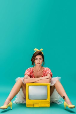Bir kadın kameraya bakarken kollarını çapraz eğip turkuazda sarı renkli televizyona yaslanıyor.