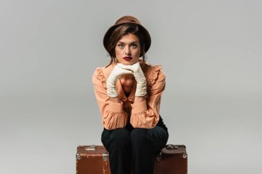 pretty, trendy woman sitting on vintage suitcase while looking at camera on grey clipart