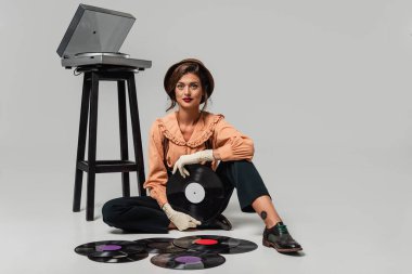 Gri pikabın ve vinil disklerin yanında oturan retro elbiseli genç kadın kameraya bakıyor.
