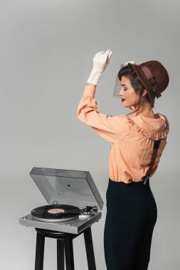 Şapkalı ve retro elbiseli modaya uygun bir kadın. Gri renkte izole edilmiş antika plak çaların yanında gözleri kapalı dans ediyor.