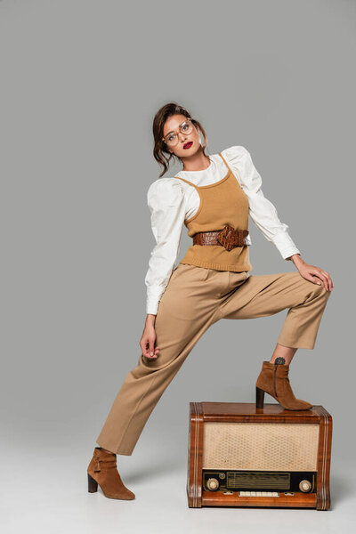 full length view of stylish woman in retro clothes stepping on vintage radio receiver on grey