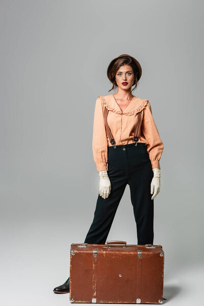 модная женщина в ретро-одежде позирует возле винтажного чемодана на сером