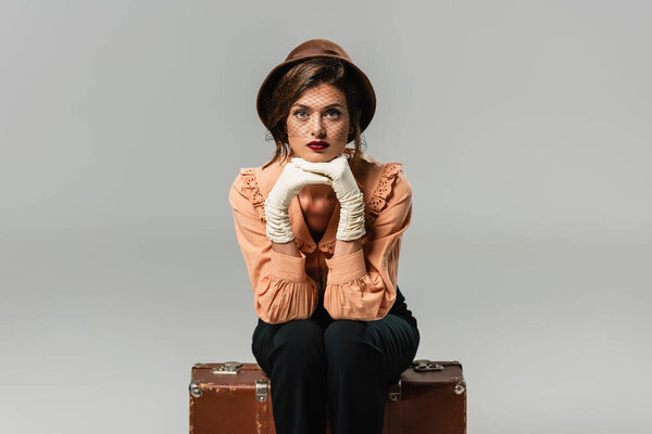 pretty, trendy woman sitting on vintage suitcase while looking at camera on grey