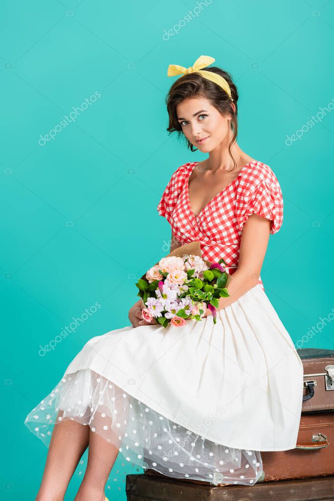 smiling woman in retro clothes sitting on vintage suitcases with flowers isolated on turquoise
