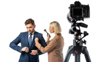 blonde makeup artist with cosmetic brush near news anchor buttoning his blazer isolated on white, blurred foreground clipart
