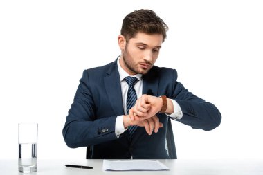 Genç haber spikeri, gazetelerin yanındaki kol saatine bakıyor ve masasındaki su bardağını bembeyaz gösteriyor.