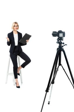 full length view of smiling news anchor on high stool pointing with hand near digital camera on tripod clipart