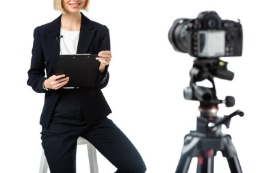 cropped view of smiling anchorwoman with clipboard sitting near digital camera on blurred foreground isolated on white clipart